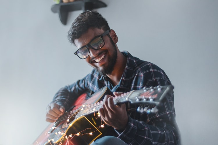 Pria yang tersenyum dan menggunakan baju kotak-kotak sedang bermain gitar.